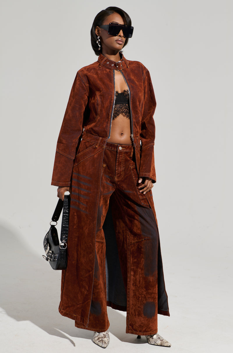 A person stands confidently, draped in the VALERIE VELVET EFFECT TRENCH paired with matching wide-legged pants, showcasing a high-low silhouette. They accessorize with oversized sunglasses, a black lace top, a studded handbag, and snakeskin-patterned boots against a light gray background.
