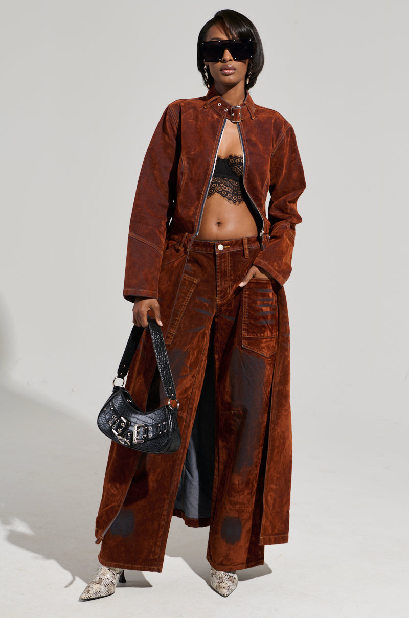 A person poses confidently in the chic VALERIE VELVET EFFECT TRENCH paired with matching pants. They sport a lacy black top, dark sunglasses, and hold a small black bag, all perfectly complemented by white pointed shoes to complete this high-low silhouette of a fashion-forward look.