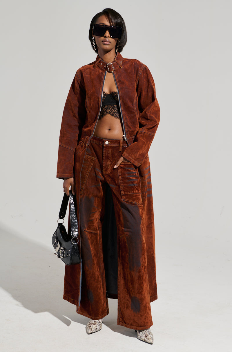 Against a plain backdrop, an individual models the VALERIE VELVET EFFECT TRENCH in a rich brown hue, beautifully styled with pants and a black lace top. Their look is enhanced by stylish sunglasses and a black handbag, adding sophistication to the high-low silhouette.