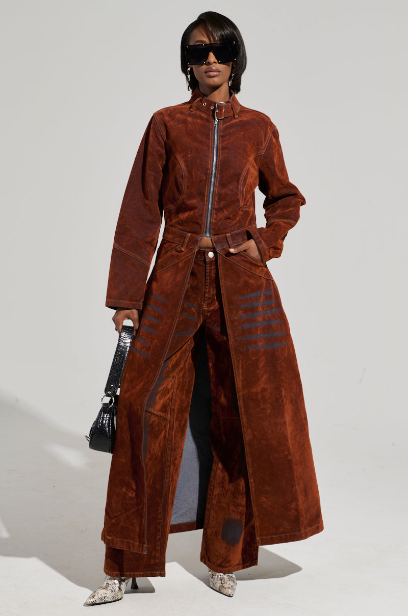 Wearing the VALERIE VELVET EFFECT TRENCH and coordinating wide-legged pants, an individual poses against a plain backdrop, showcasing a chic high-low silhouette. They accessorize with a small black handbag, dark sunglasses, and snakeskin ankle boots. Their hair is styled in a sleek bob.
