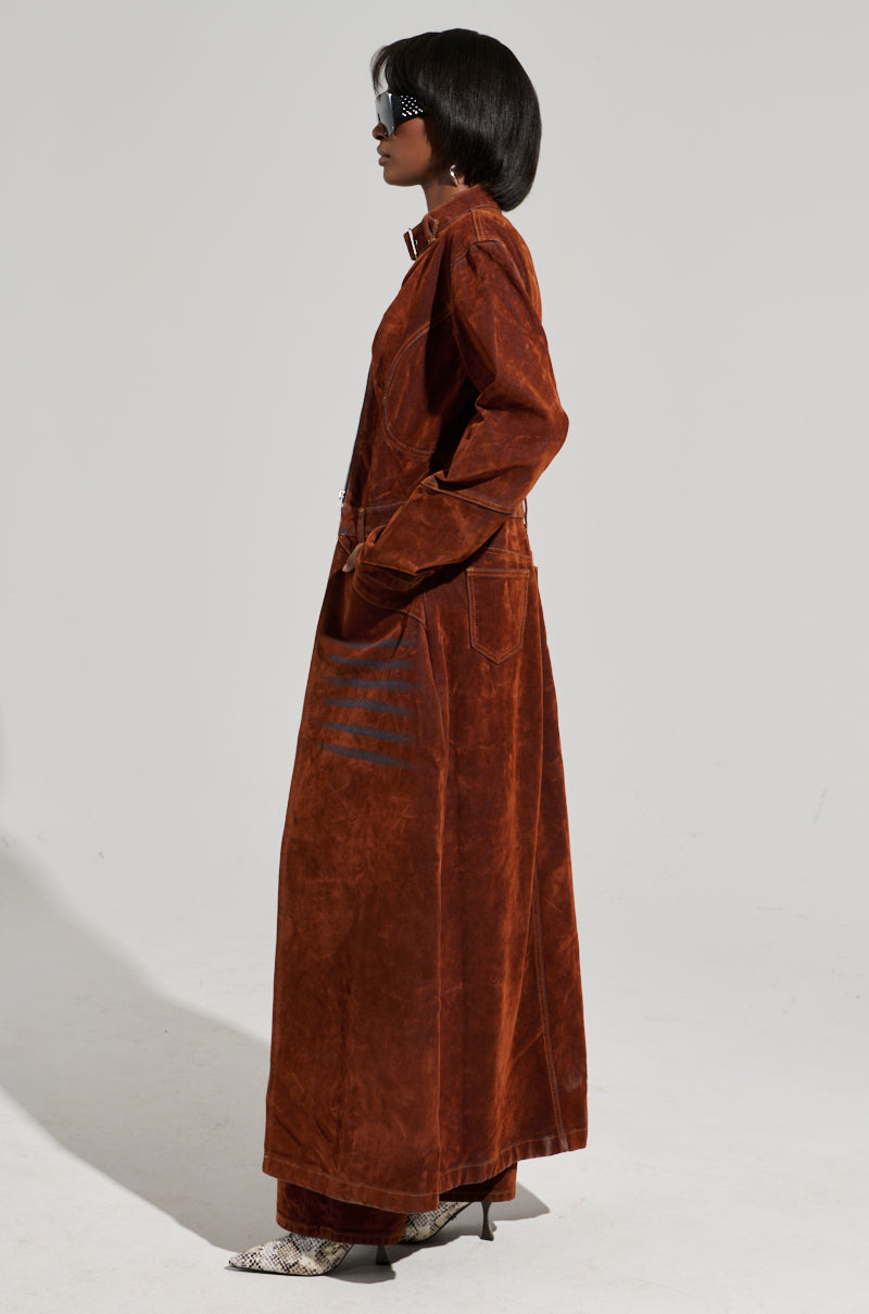 A person wearing the VALERIE VELVET EFFECT TRENCH at maxi length and patterned boots stands in profile against a plain background. They have short dark hair and are accessorized with black sunglasses.