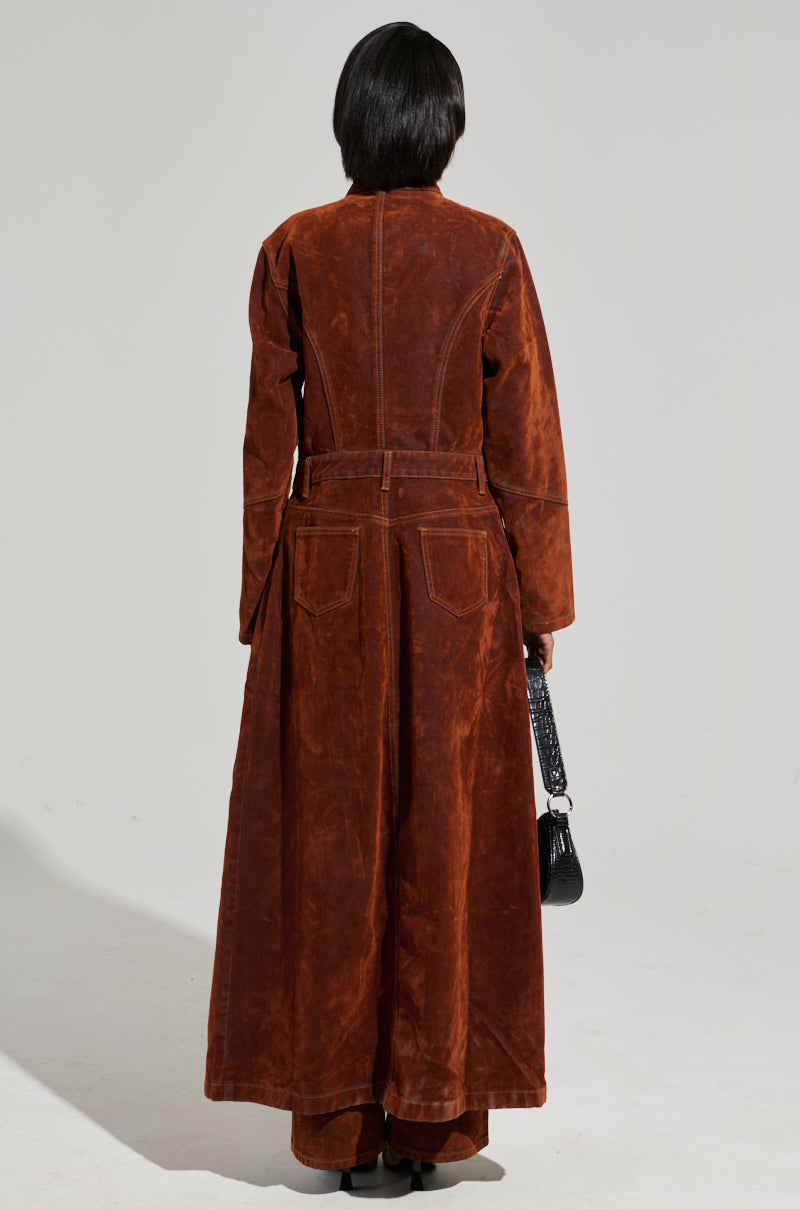 A rear view of someone wearing the VALERIE VELVET EFFECT TRENCH, a long suede coat in brown with prominent seams and stitching details, showcasing a high-low silhouette. They are holding a small black handbag against a plain background.