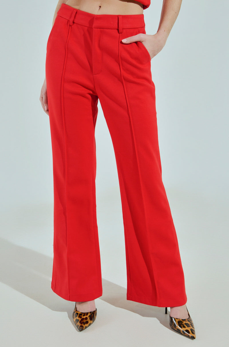 The woman showcases the Business Casual Trouser in Red, featuring high-waisted, wide-leg flair, front pleats, and a belt. Paired with leopard print heels for a chic professional look, she confidently rests one hand in her pocket.