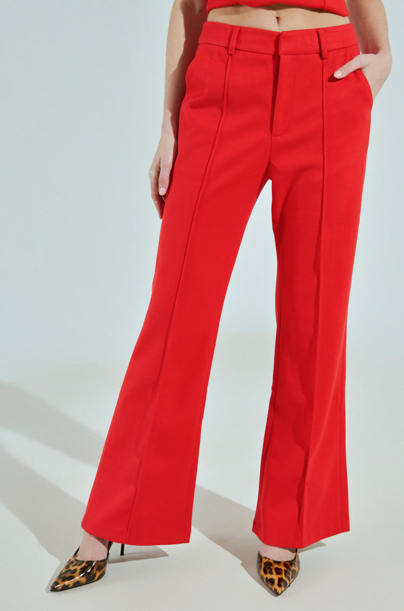 Standing against a light-toned backdrop, the individual wears BUSINESS CASUAL TROUSER IN RED, showcasing professional chic with high-waisted, bright red flared pants. Their leopard print heels peek out as their arms rest calmly at their sides, complementing the sophisticated attire.