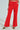 Standing against a light-toned backdrop, the individual wears BUSINESS CASUAL TROUSER IN RED, showcasing professional chic with high-waisted, bright red flared pants. Their leopard print heels peek out as their arms rest calmly at their sides, complementing the sophisticated attire.