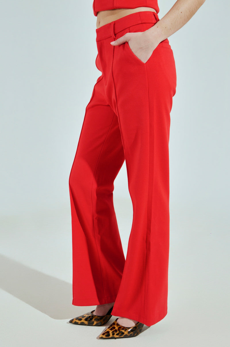 A person models vibrant BUSINESS CASUAL TROUSER IN RED, complete with a matching belt and hands in pockets. Paired with pointed leopard print shoes, the look stands out against a plain light backdrop, emphasizing the chic and professional style.