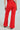 Rear view of a person wearing BUSINESS CASUAL TROUSER IN RED paired with a matching top, epitomizing professional chic. They stand with one hand outstretched. The high-waisted flared pants have belt loops and welt pockets, with glimpses of brown and black-patterned shoes visible.
