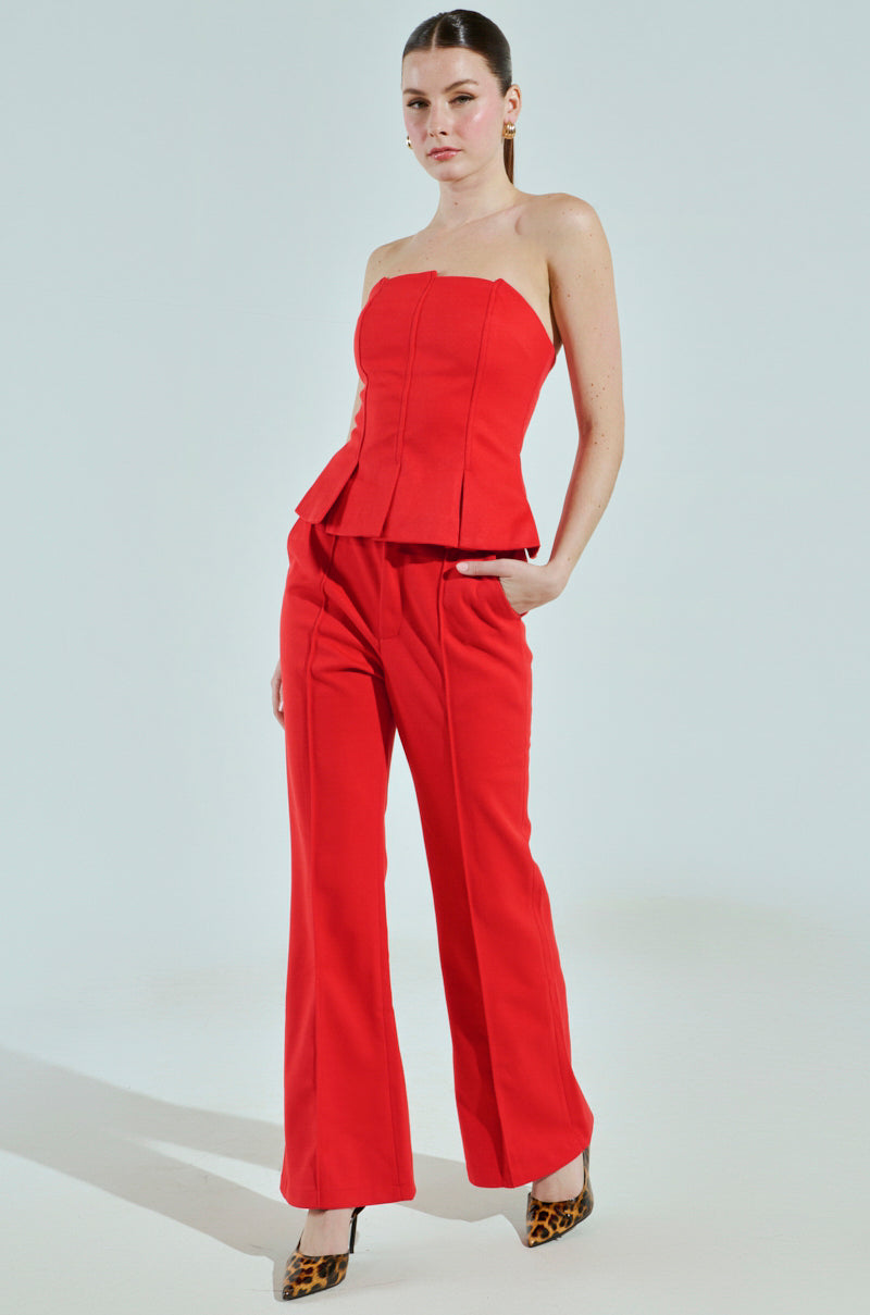 A person confidently models the red BUSINESS CASUAL CORSET TOP from AKIRA Label with coordinating wide-leg pants. One hand in a pocket, they pair it with leopard print heels against a plain light background, embodying edgy business casual chic.