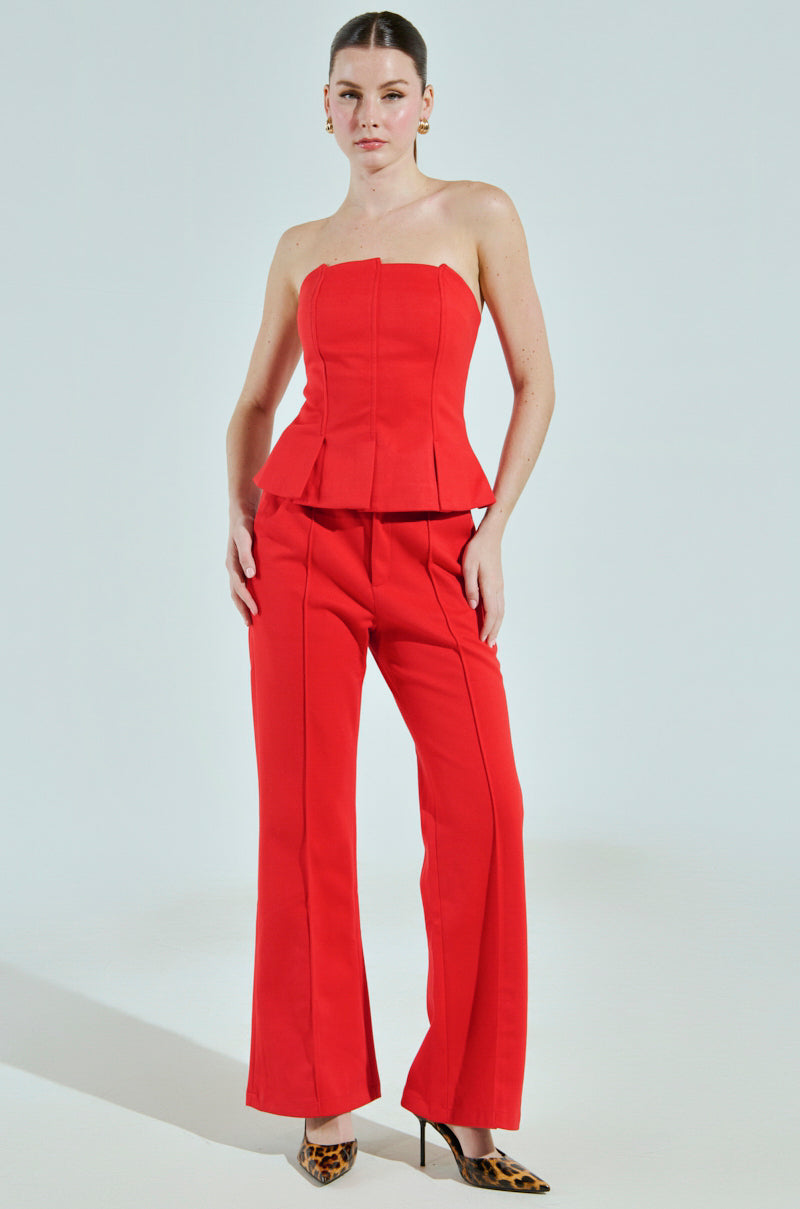 A woman sports the AKIRA Label's CHIC BUSINESS CASUAL CORSET TOP IN RED, paired with flared pants and leopard print high heels, set against a plain, light-colored background.