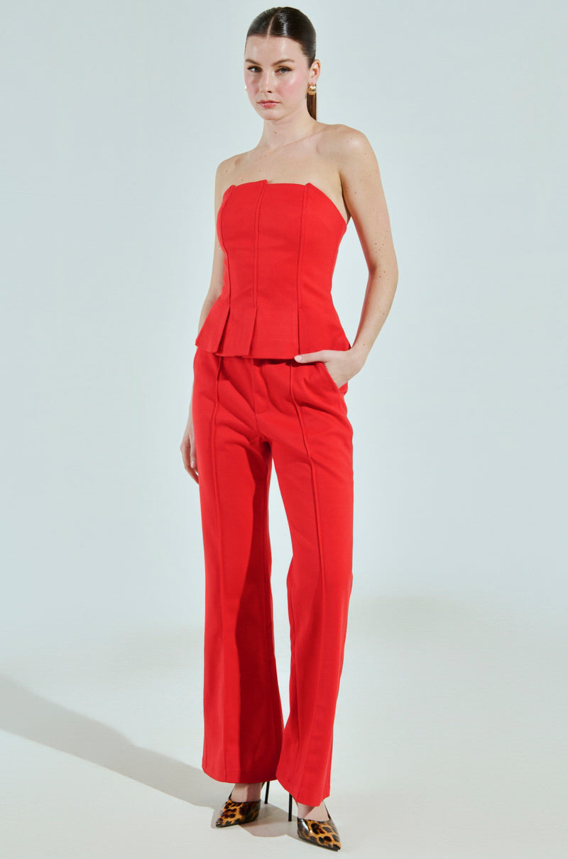 Dressed in a BUSINESS CASUAL CORSET TOP IN RED, along with matching wide-leg red pants, and animal-print high heel shoes from the AKIRA Label collection, an individual strikes a pose against a plain backdrop with one hand in their pocket.