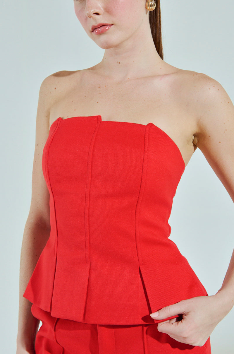 A person with light skin wears a red strapless, fitted BUSINESS CASUAL CORSET TOP IN RED by AKIRA Label, featuring pleats on the front and structured fabric. Gold stud earrings complement the look against a plain, light-colored background adding elegance.