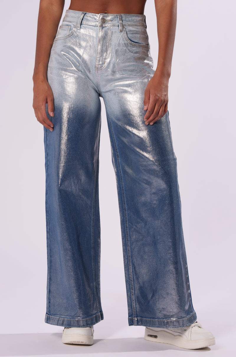 A person wearing high-rise waist, wide-leg LITTLE MISS SUNSHINE DENIM PANT in metallic silver, paired with white sneakers. The pants boast a metallic sheen denim look, blending a shiny texture with a denim-blue color. The background is plain white.