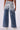 An individual is depicted from behind wearing the LITTLE MISS SUNSHINE DENIM PANT, which boasts a shimmering metallic blue and silver ombré design. These high-waisted, wide-leg jeans feature two back pockets and are styled with white sneakers against a simple white backdrop.