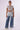 A person stands against a white background, dressed in a sheer, short-sleeve crop top with a mesh design and high-waisted LITTLE MISS SUNSHINE DENIM PANT featuring a metallic sheen, paired with white sneakers. They are accessorized with a silver necklace.