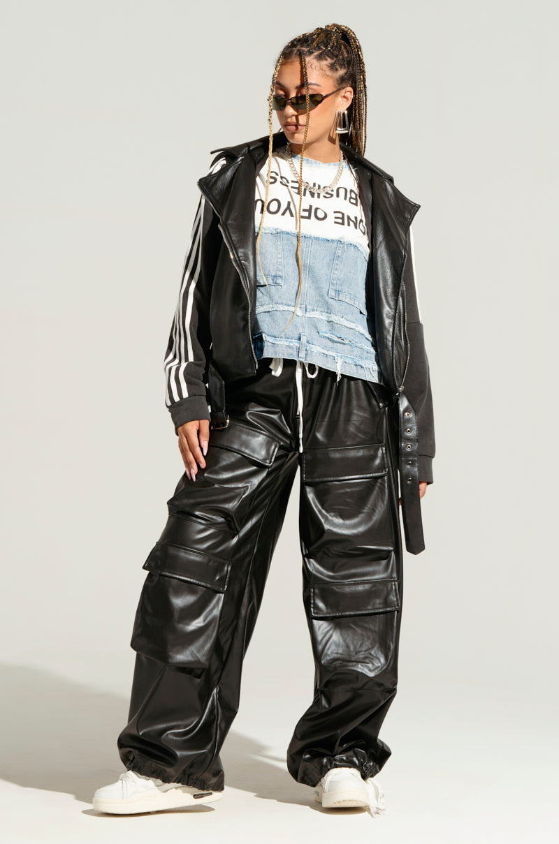 A person with sunglasses and a braided hairstyle stands confidently in a black leather jacket and RICK WIDE LEG FAUX LEATHER PANTS, layered over a denim vest and a graphic tee with white sneakers. Cargo pockets add an edgy touch to their style against the neutral gray background.