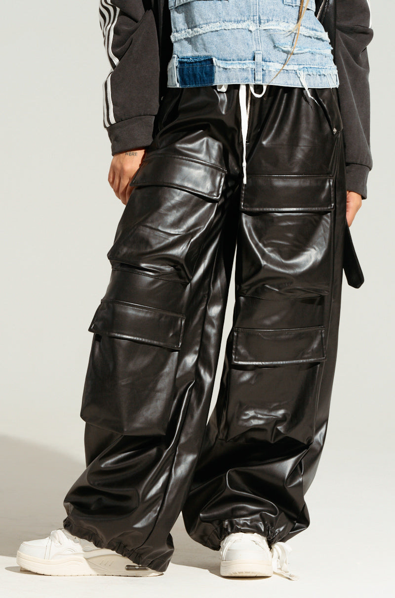 A person dons a black and blue patchwork top combined with the RICK WIDE LEG FAUX LEATHER PANT, which features cargo pockets, presenting a casual streetwear vibe. Their hands rest relaxed by their sides, while white sneakers effortlessly complete the cool look.