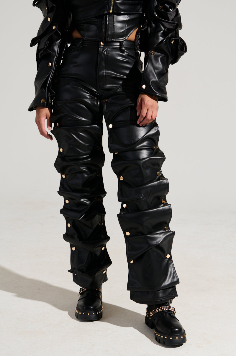 A person sporting the SNAP OUT OF IT FAUX LEATHER PANT in black, characterized by ruched, high-rise design adorned with gold studs. They paired it with studded black boots. The ensemble is completed with dramatic, voluminous sleeves and snap-button removable paneling. 0