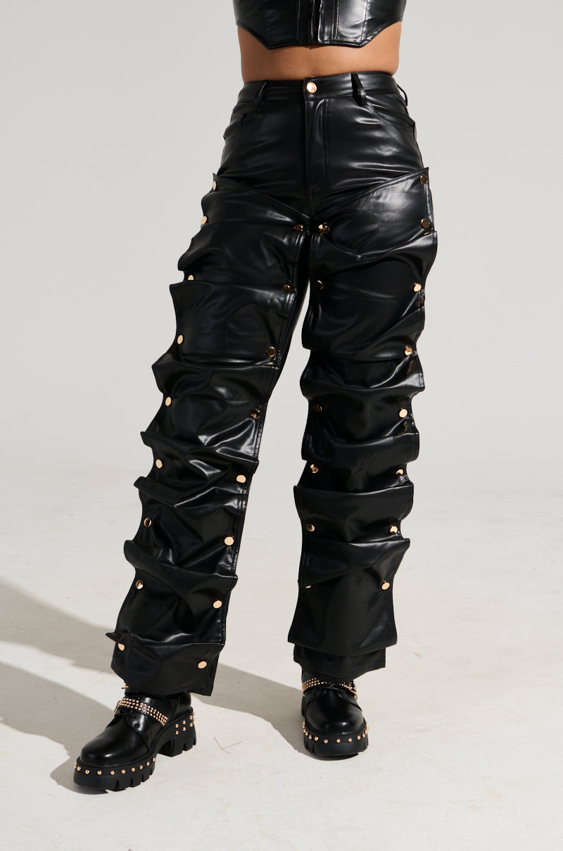 A person wearing the SNAP OUT OF IT FAUX LEATHER PANT, which are high-waisted with ruched detailing and gold studs, paired with black chunky boots adorned with gold chains. The background is plain, and the focus is on the lower half of the person’s body. 2