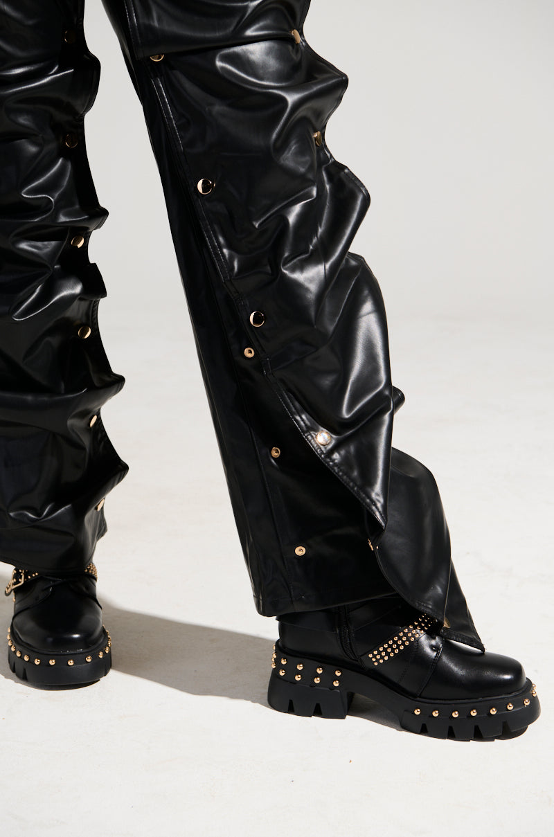 A person wearing the SNAP OUT OF IT FAUX LEATHER PANT, which features high rise, gold eyelet detailing, and a distinct ruched design for a textured appearance. They are paired with matching black chunky studded shoes. The image highlights the lower half of the outfit against a plain background. 8