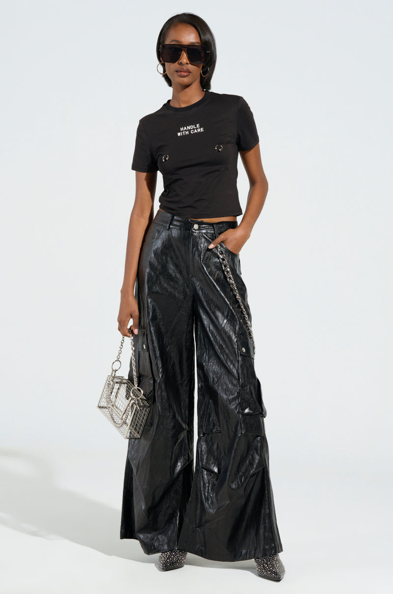 Dressed in a black t-shirt featuring the text "Handle With Care," they pair it with SOPHIE FAUX LEATHER TROUSER, styled in a high-rise, wide-leg silhouette. Adorning sunglasses and gripping a small metallic handbag, they complete the look with patterned shoes while posing confidently against a plain background.