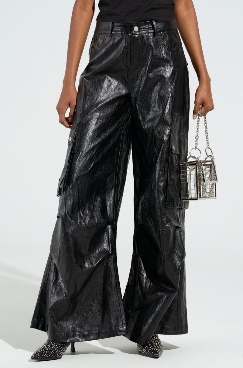 A person is wearing the SOPHIE FAUX LEATHER TROUSER, featuring a wide-leg and high-rise design, while holding a small silver cage-style handbag. They stand against a plain background, donning pointed black shoes with studded details.