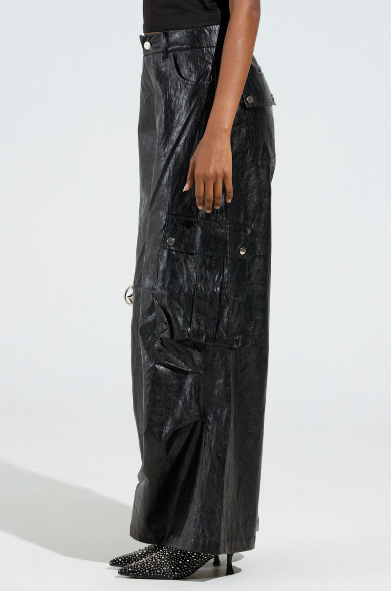 A person wearing the SOPHIE FAUX LEATHER TROUSER, which features a high-waisted, textured wide-leg design with flap pockets and buttons on the sides. The ensemble is complemented by a high rise element paired with a black top and pointed, studded black boots.
