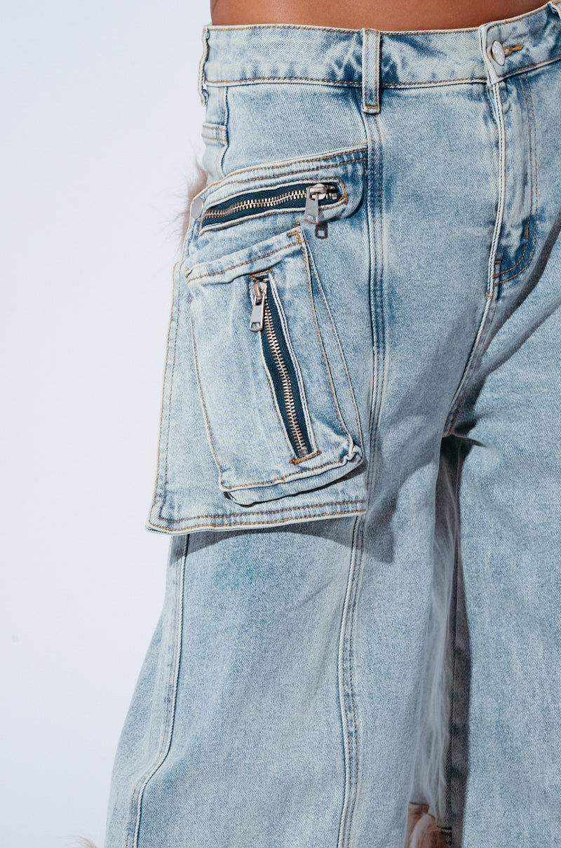 A close-up view of light-washed FUR DENIM CARGO PANTS with multiple zippers. These high-rise pants showcase a unique design featuring a large, square, zippered pocket on the side of the thigh and additional zippers on the main pocket. The denim has a slightly worn, vintage look.