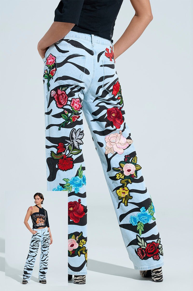 A model showcases the SLEEPING WITH ROSES DENIM PANT, featuring bold zebra print and vibrant floral embroidery with striking red, pink, and blue rose patches. Paired with a black sleeveless top, this standout ensemble is set against a plain background.