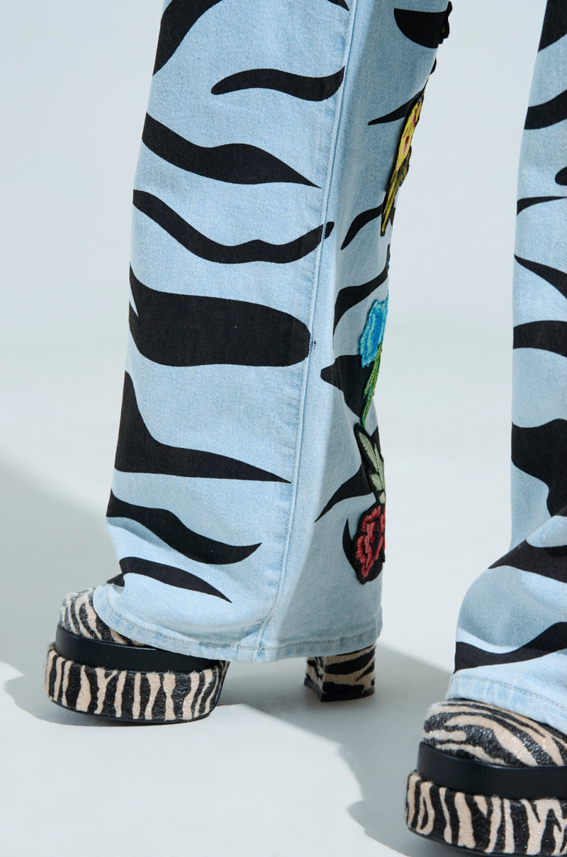 Close-up of a person in SLEEPING WITH ROSES DENIM PANT featuring black zebra stripes and vibrant rose patches, paired with platform sandals echoing the zebra pattern, against a soft, neutral background.