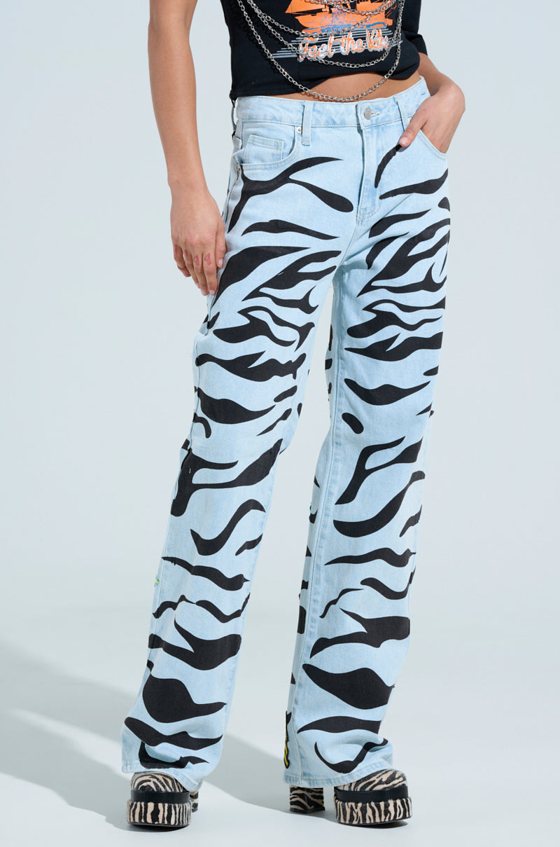 A person wears striking SLEEPING WITH ROSES DENIM PANT in light blue with bold black zebra print, paired with a sleek black top and black-and-white striped shoes. Chain accessories add edge against a plain white background.