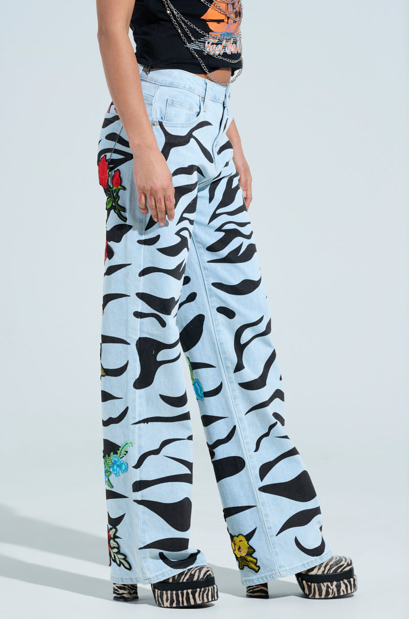 A person wears the SLEEPING WITH ROSES DENIM PANT in light blue, featuring bold black waves, floral designs, and rose patches. They're styled with a black graphic T-shirt and zebra-print platform shoes against a light gray background.