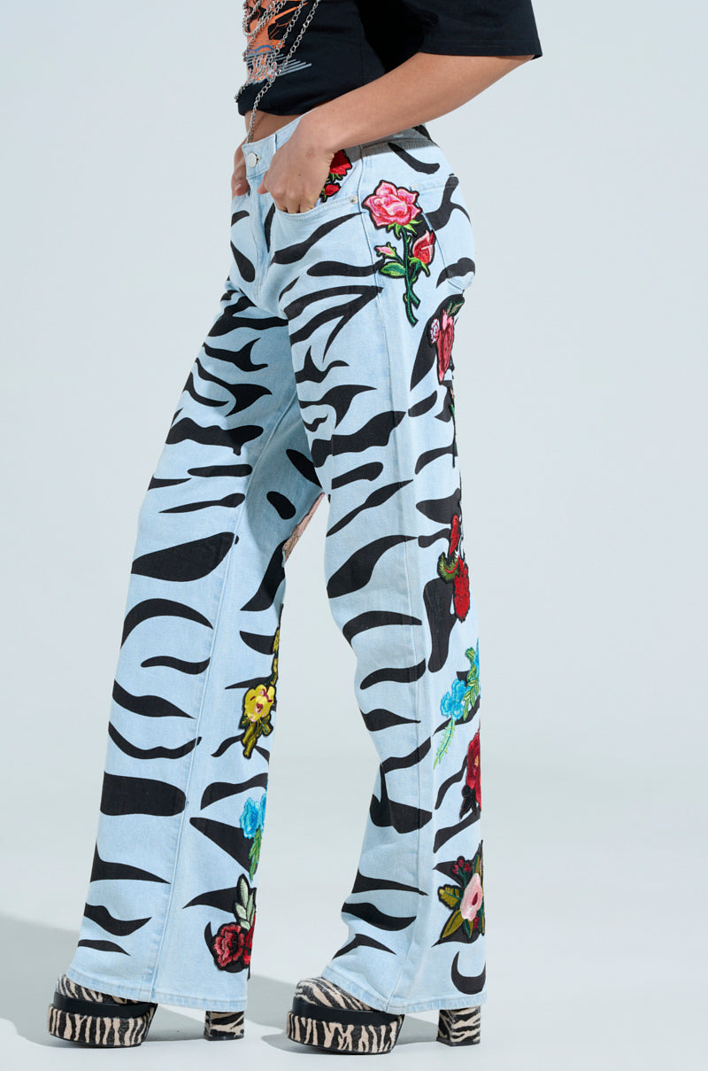 Someone wears the SLEEPING WITH ROSES DENIM PANT with black zebra stripes and rose patches, complemented by platform shoes with a similar zebra print. The upper part of their body is partially visible against a plain background.