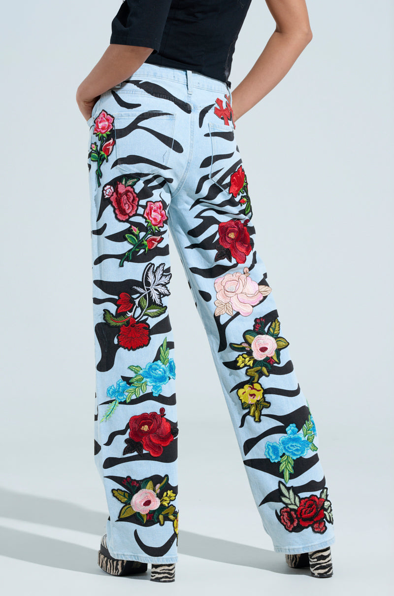 A person wears the SLEEPING WITH ROSES DENIM PANT, featuring zebra-print and vibrant floral embroidery in red, pink, blue, and yellow. Paired with a black top and zebra-patterned shoes, they stand against a plain background.