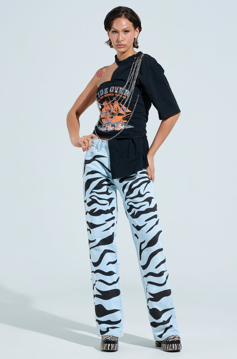 With a confident pose against a light background, they wear a one-shoulder black top adorned with an orange graphic design, paired with SLEEPING WITH ROSES DENIM PANT featuring light blue zebra stripes and patterned platform shoes. They strike an assertive stance, hand on hip and expression serious.