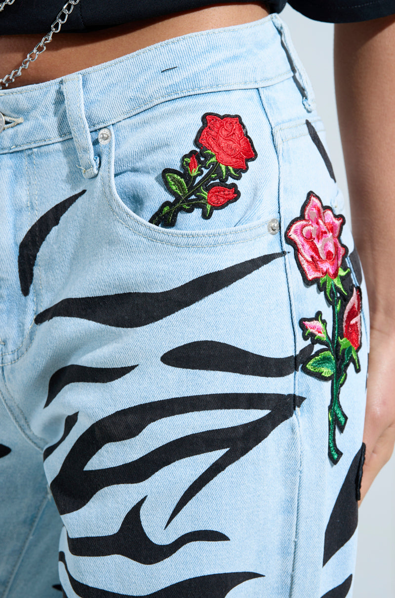 Close-up of light blue "SLEEPING WITH ROSES DENIM PANT" with black zebra print and red rose embroidery. Vibrant rose patches on the front pocket and thigh create a colorful contrast to the striking zebra pattern.