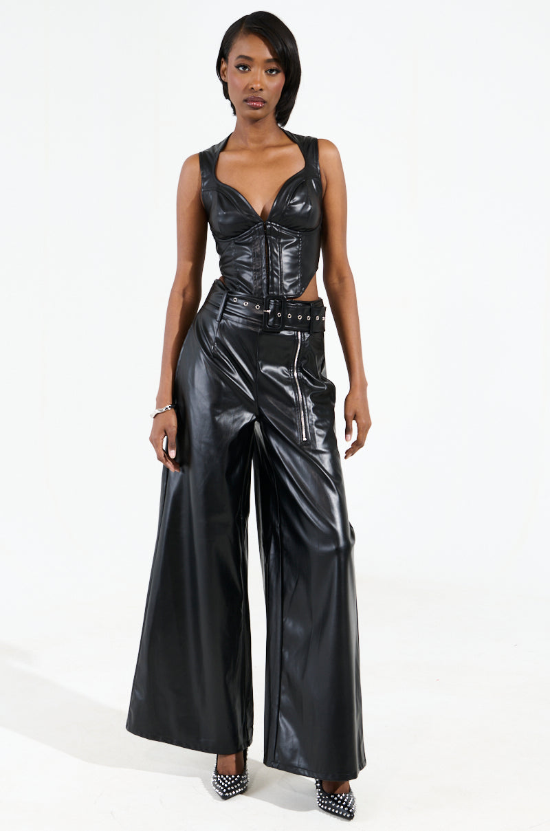 A person stands confidently in a black leather outfit, featuring a sleeveless top and SHAINA FAUX LEATHER TROUSER with a detachable belt. With short hair and black studded heels, they create a striking silhouette against the plain white background.