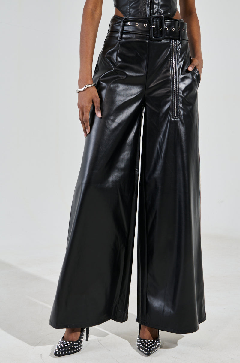 A person wears SHAINA FAUX LEATHER TROUSERS, featuring a high-rise wide-leg fit in black faux leather with a detachable belt, hands casually resting in pockets. They pair these with studded high-heeled shoes against a plain backdrop, letting the chic outfit shine.