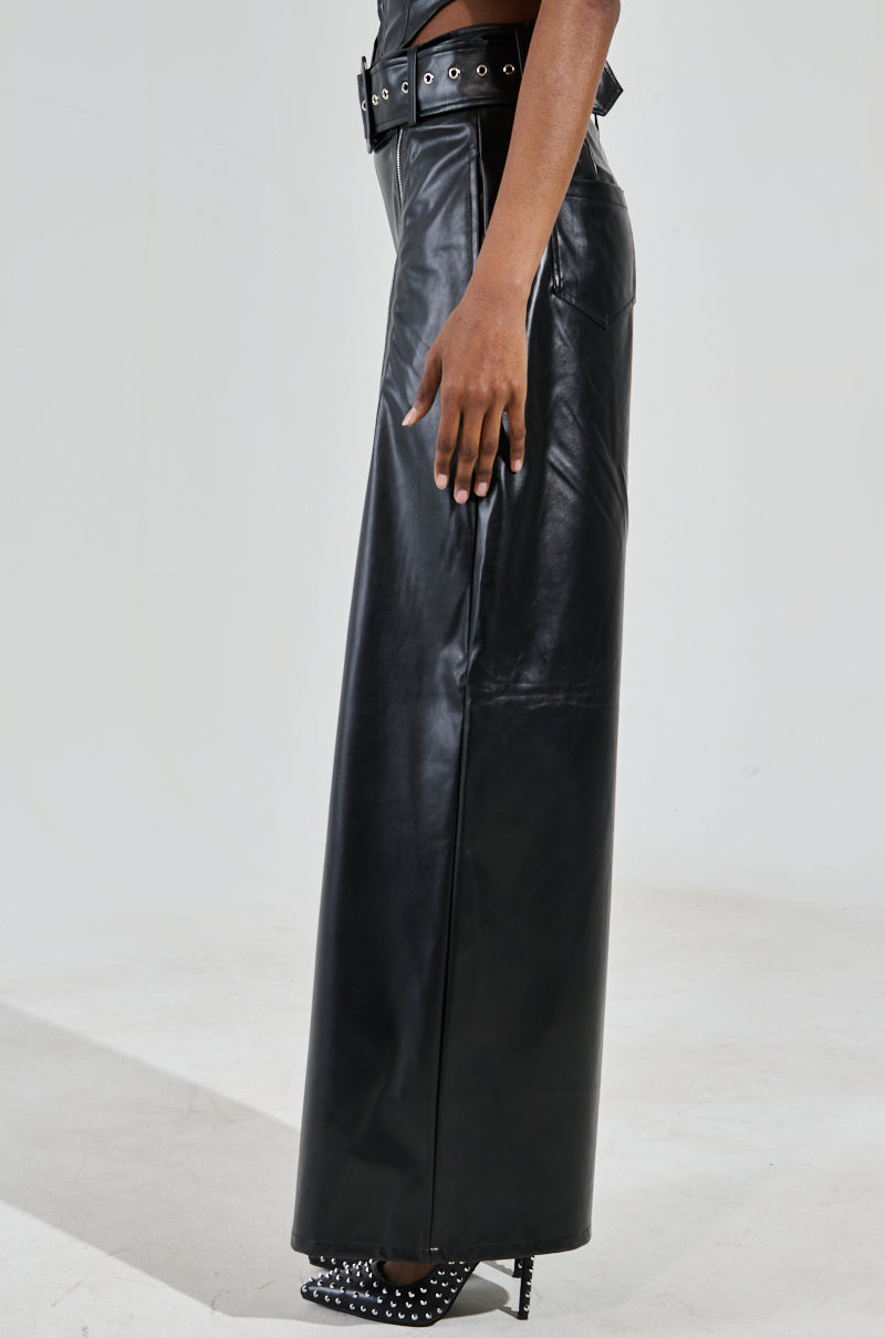 Wearing the SHAINA FAUX LEATHER TROUSER with a detachable belt, someone stands on a light surface, paired with black studded high heels. Only their lower half is visible.