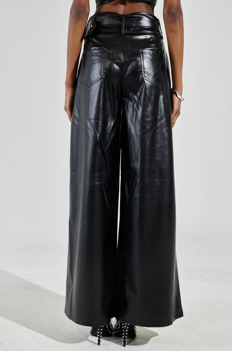 Rear view of a person wearing SHAINA FAUX LEATHER TROUSER—high-rise, wide-leg with pockets and glossy finish. The pants include a detachable belt, complemented by heels featuring spiked details for a chic look.
