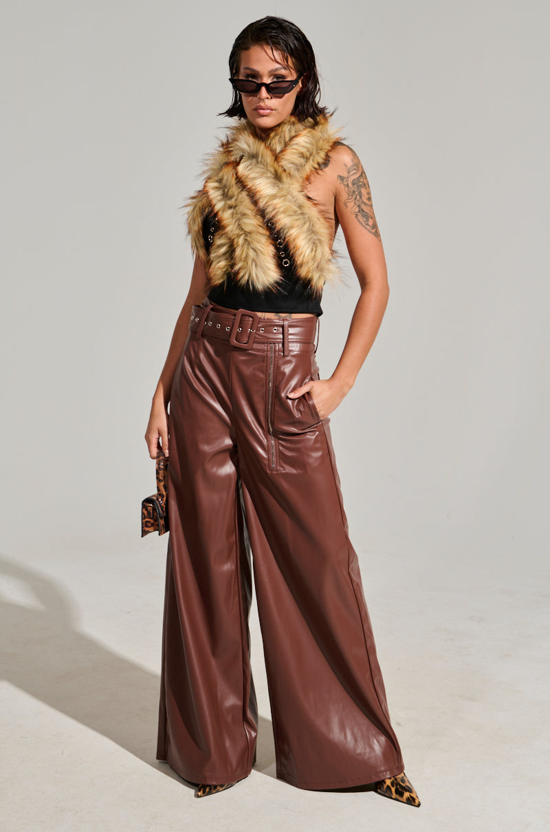 A person poses confidently in a chic ensemble that includes a fur scarf, sleeveless top, and the SHAINA FAUX LEATHER TROUSER in a high-rise brown design. Complementing the look with sunglasses and a small purse, they stand against a neutral-toned backdrop.