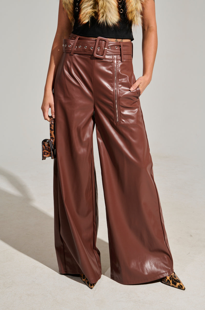 A person showcases the SHAINA FAUX LEATHER TROUSER, a high-rise design in brown with wide-leg cuts, a matching belt, and convenient side pockets. This chic look is complemented by a small leopard-print purse and a furry top that pairs seamlessly with leopard-print shoes. A neutral background highlights this stylish ensemble.