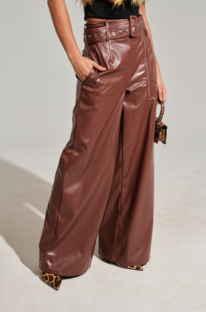 A person is dressed in SHAINA FAUX LEATHER TROUSERS, which feature a high-rise silhouette and a brown faux leather finish, complete with a matching belt. Complementing the look is a small animal print handbag and leopard print shoes. The outfit's wide-leg cut stands out beautifully against the plain, neutral backdrop.