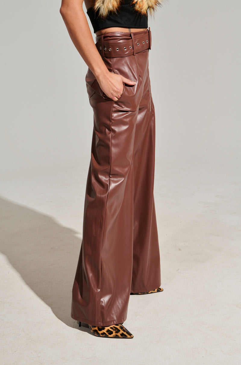A person stands on a light-colored floor wearing the SHAINA FAUX LEATHER TROUSER, which features a high waist and wide belt. With their left hand resting in the pocket, they pair the trousers with pointed leopard-print shoes. The top they are wearing has partially visible fur details.