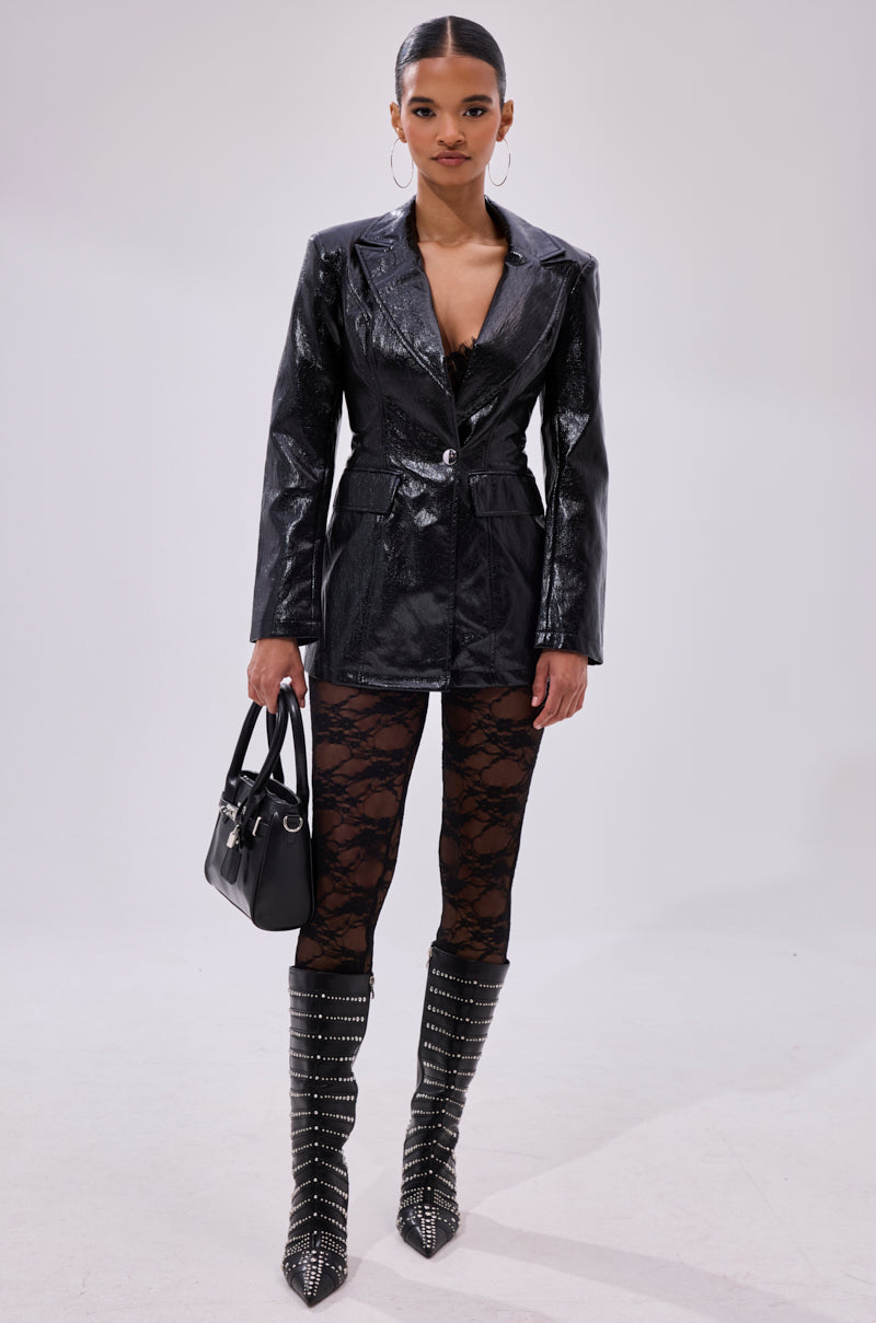 A woman poses against a plain background wearing FEEL FREE TO UNZIP ME LACE LEGGING, a black faux leather blazer, studded knee-high boots, hoop earrings, and holding a black handbag.