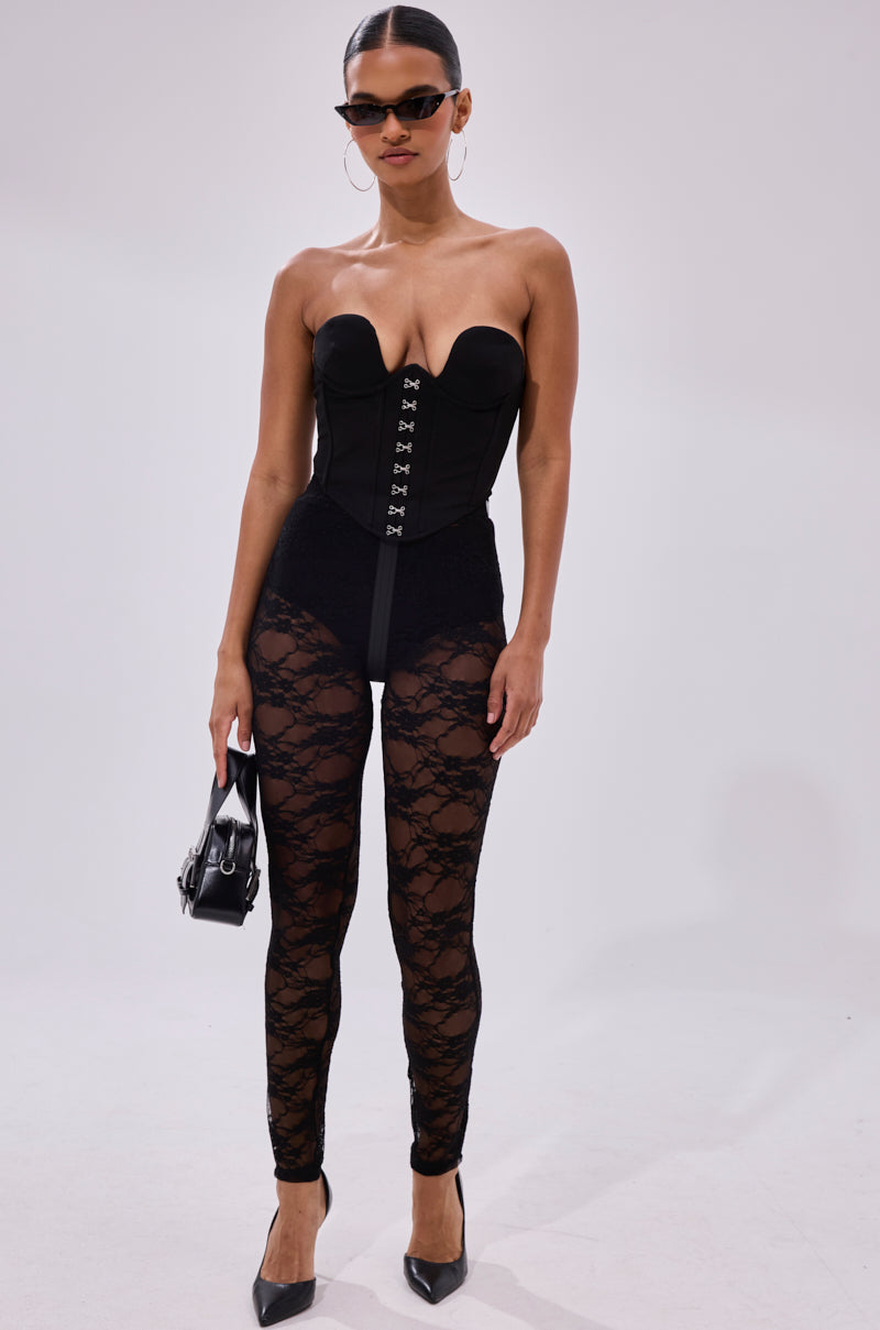 A woman poses against a plain background wearing FEEL FREE TO UNZIP ME LACE LEGGING, a strapless black corset, pointed black heels, black sunglasses, and holding a small black handbag.