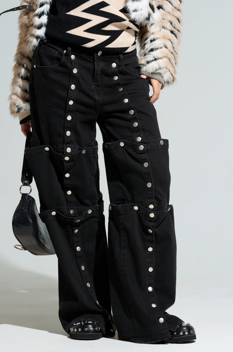 A person wearing the Snap Out of It Denim Pant in black, featuring a high-rise wide-leg cut with a snap button design, paired with a patterned black and white sweater and an elegant fur coat. They are holding a small black handbag and sporting studded black shoes.