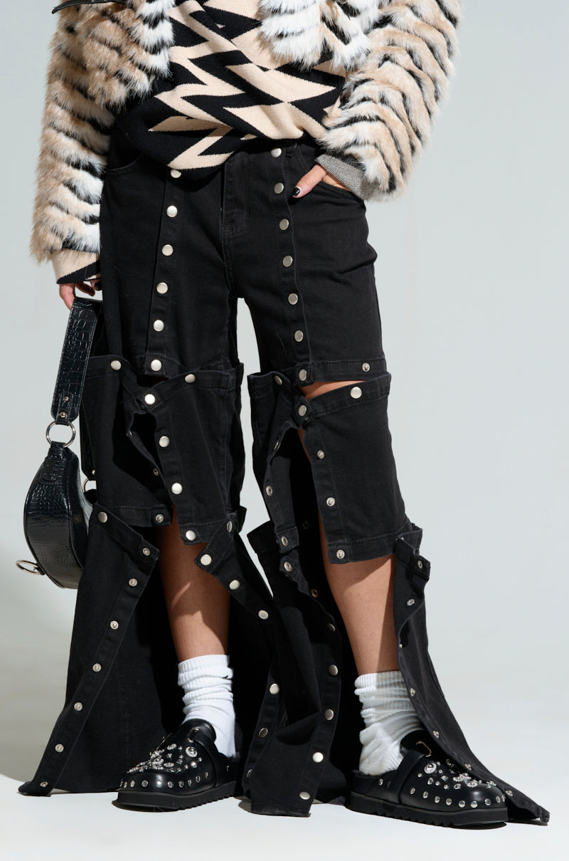 A person is dressed in the SNAP OUT OF IT DENIM PANT IN BLACK, featuring an edgy snap button design and high-rise wide-leg silhouette. They're also wearing a black and white zig-zag patterned top, complemented by black studded shoes and white socks, while carrying a black handbag.
