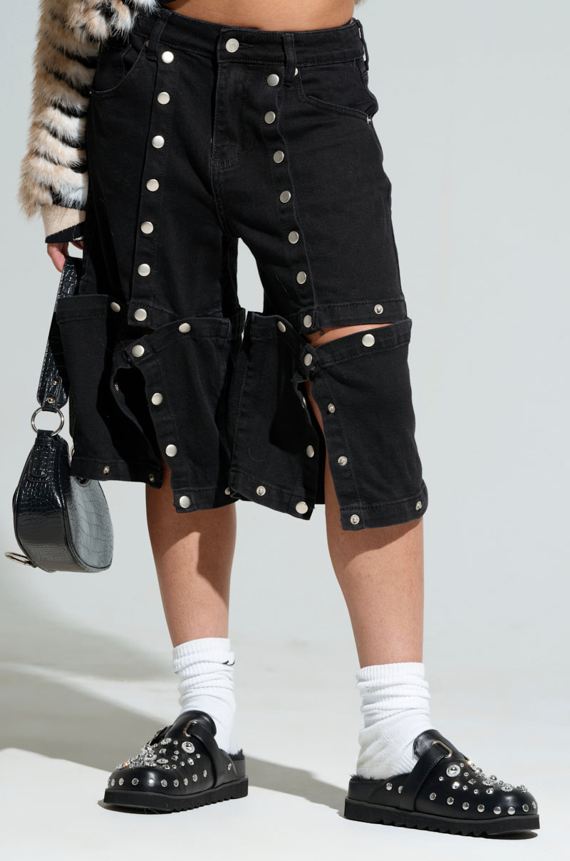 A person wearing the "Snap Out of It" denim pants in black, featuring metal studs and cut-out details, showcases their knee-length style. Their high-rise wide-leg silhouette perfectly coordinates with snap button design shoes and white socks, while they carry a textured black handbag. A patterned coat drapes over their shoulder for a hint of added style.