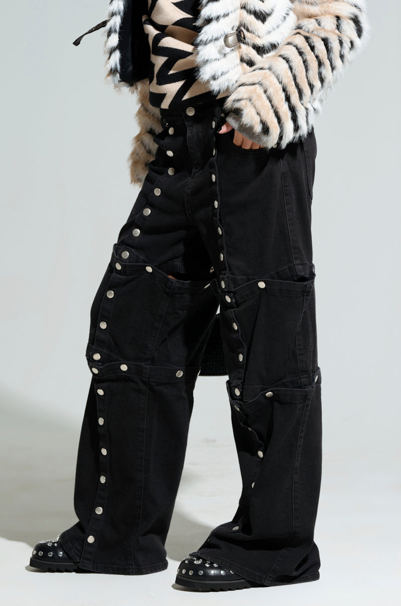 A person wearing the SNAP OUT OF IT denim pant in black, featuring a high-rise wide-leg design with multiple silver buttons and geometric stitching. The pants are paired with a black and white fur jacket and studded black shoes. Their upper face and background remain out of view.