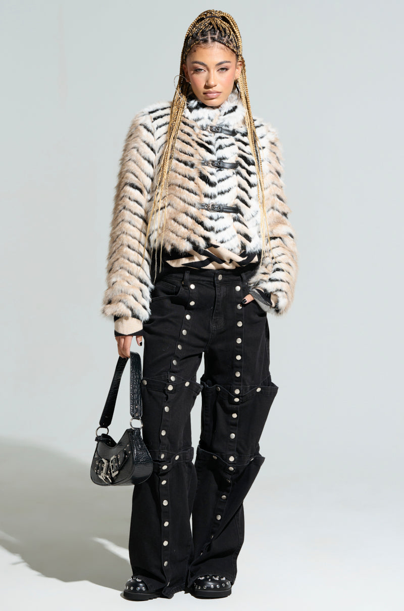 A person with braided hair stands confidently against a neutral background, wearing a striped fur jacket and the "Snap Out of It" denim pants in black, featuring a high-rise wide-leg cut adorned with snap button design. They hold a small black bag, completing a bold and stylish look.